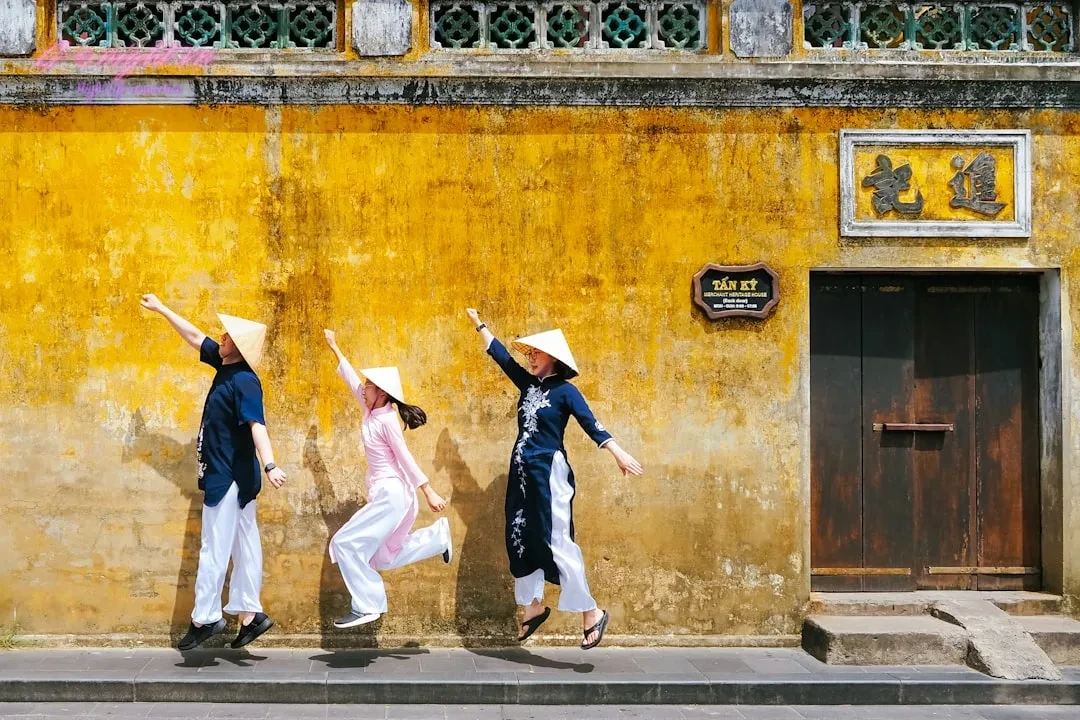 Three people in conical hats jump against yellow wall